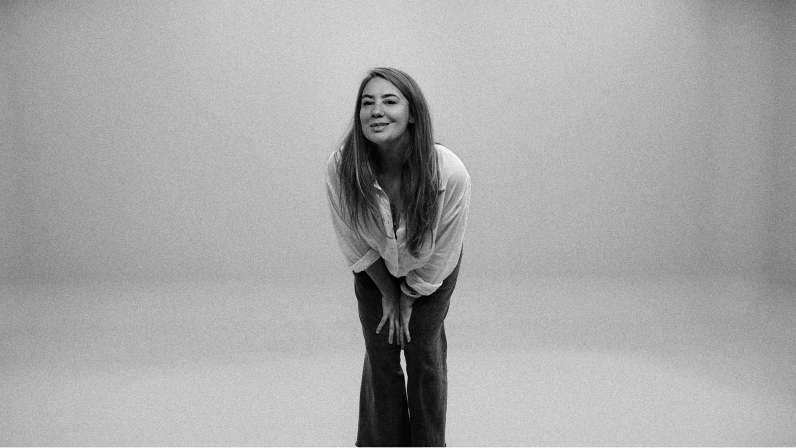Margot Dandieu, en chemise blanche, penchée en avant et souriante, sur fond clair.