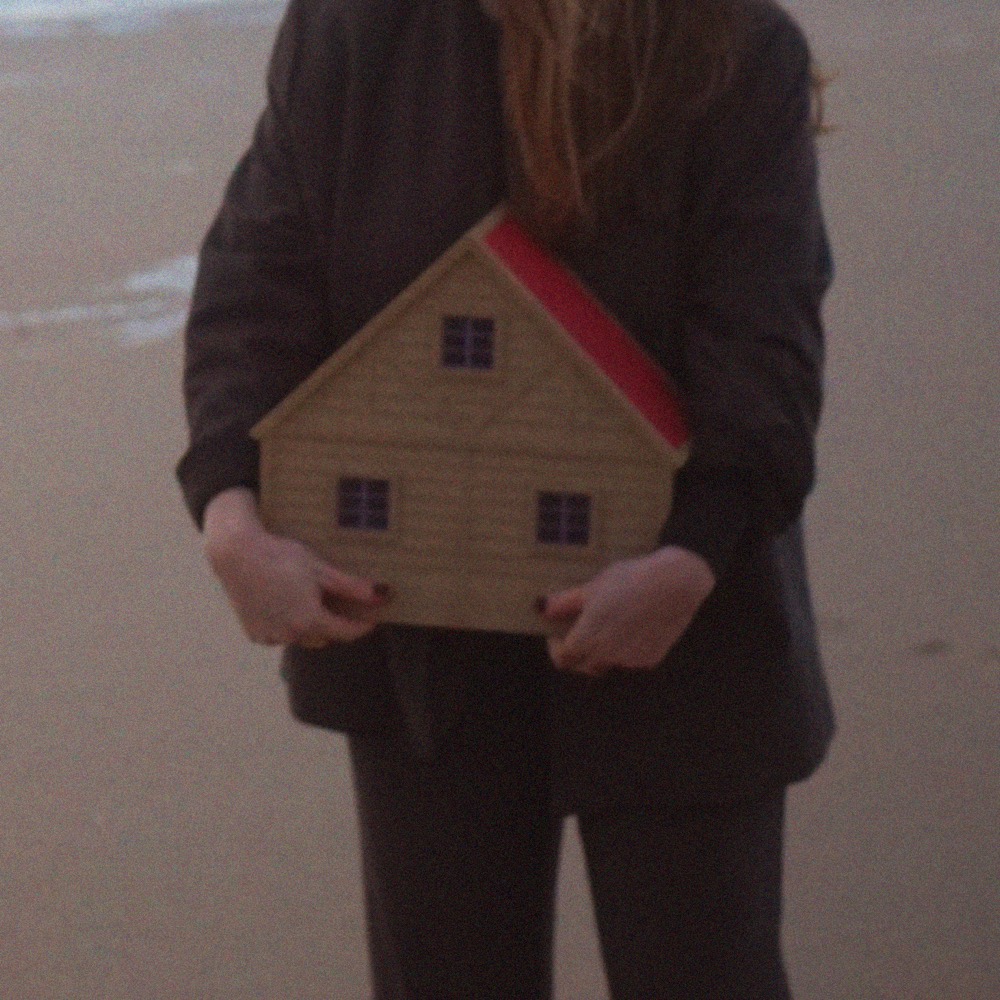 Buste de Margot Dandieu en manteau sombre, tenant à deux mains une maquette de maison en bois à toit rouge, sur une plage.