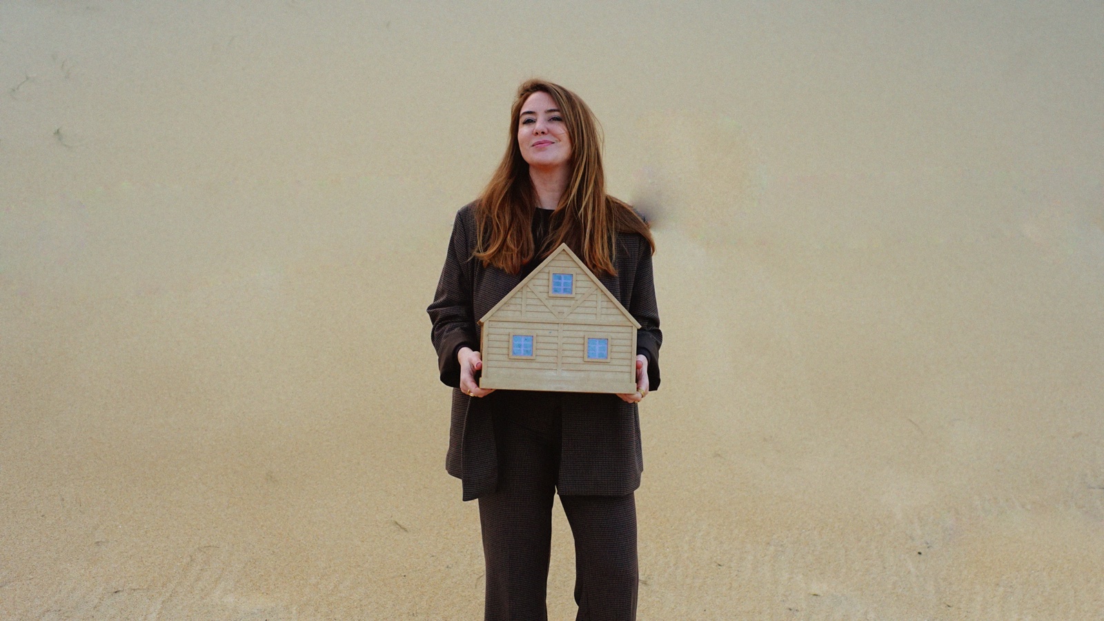 Margot Dandieu, en manteau sombre, tenant à deux mains une maquette de maison en bois devant un mur couleur sable.